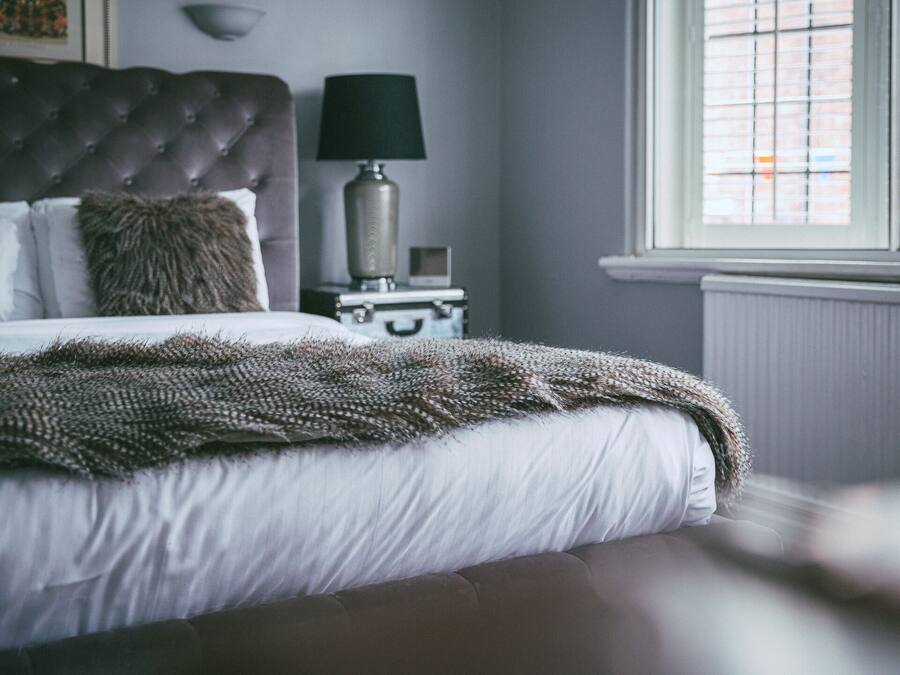Un dormitorio con cabecera capitonada, lámpara de noche, maleta y ventana con persianas.
