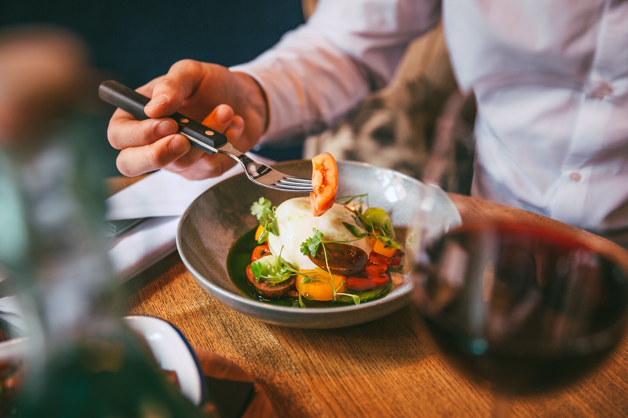 A person dining at a table with a bowl of food, a fork, and a glass of red wine.