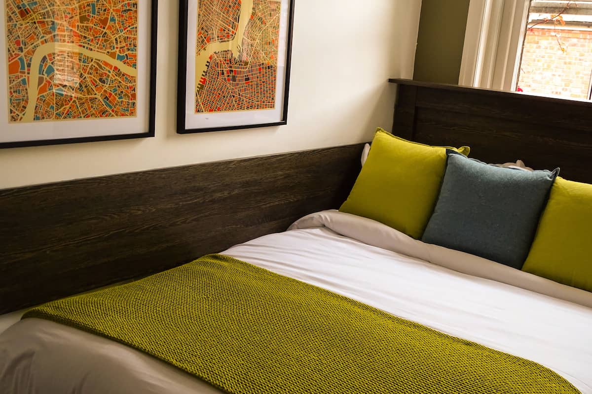 Un letto rifatto con cura, con una coperta verde e cuscini giallo, blu e grigio. Due opere d'arte raffiguranti paesaggi urbani sono appese alla parete sopra il letto.