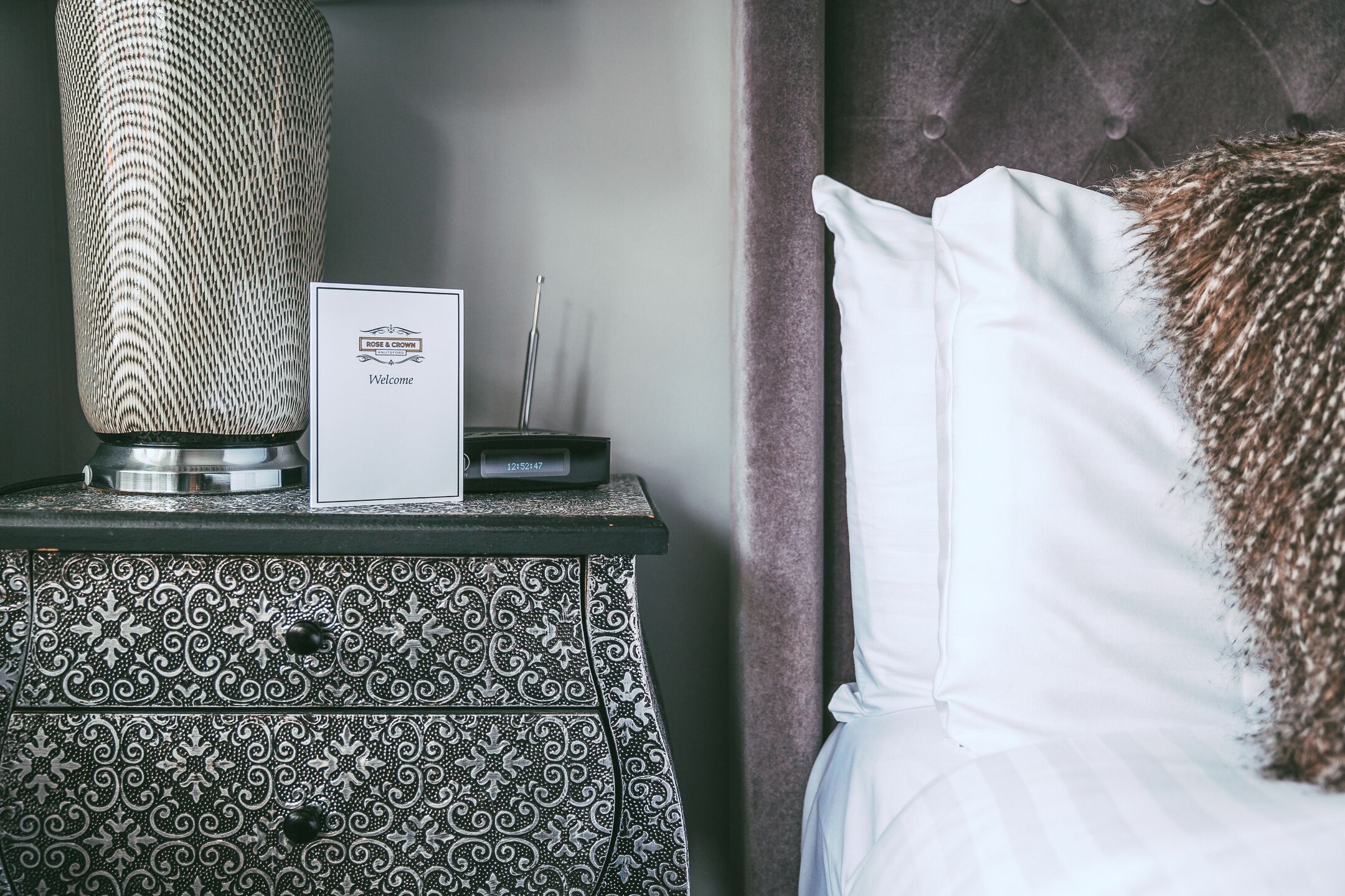 A neatly made bed with white linens and a decorative pillow, a patterned vase, and a framed welcome sign on a bedside table.