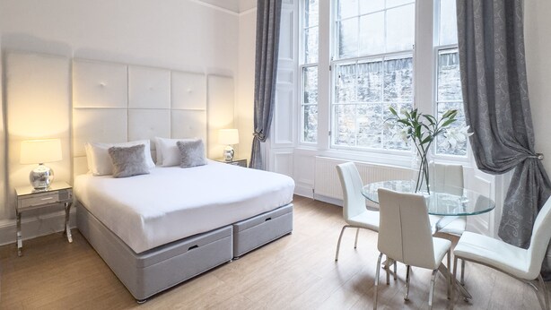A modern hotel room with a large bed, bedside tables, a glass table with chairs, and a view of the city through large windows. - Hanover 71 Suites (Edinburgh)