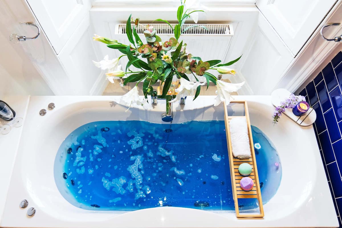 Un baño con una bañera llena de agua azul, una bandeja de madera con productos de baño y un jarrón con flores.