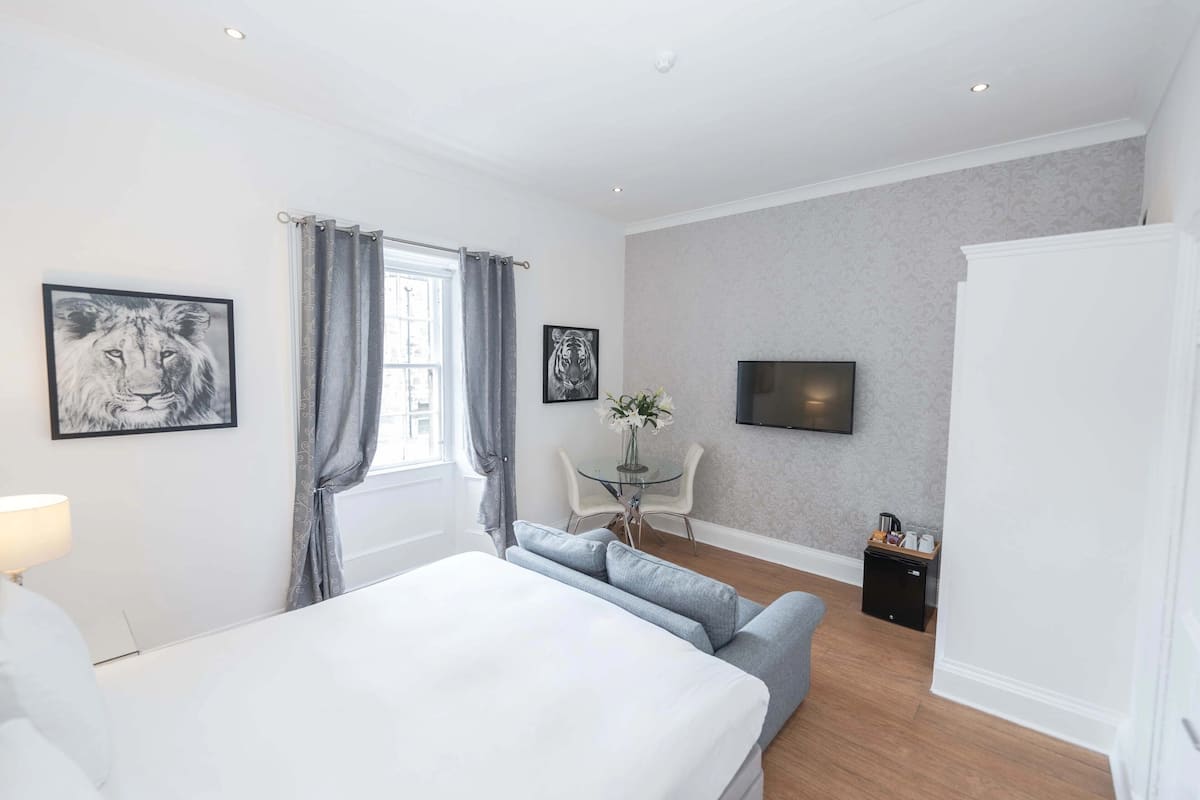 Una habitación de hotel moderna con una cama grande, un área de descanso con una mesita y sillas, un televisor de pantalla plana en la pared y un refrigerador en una esquina.