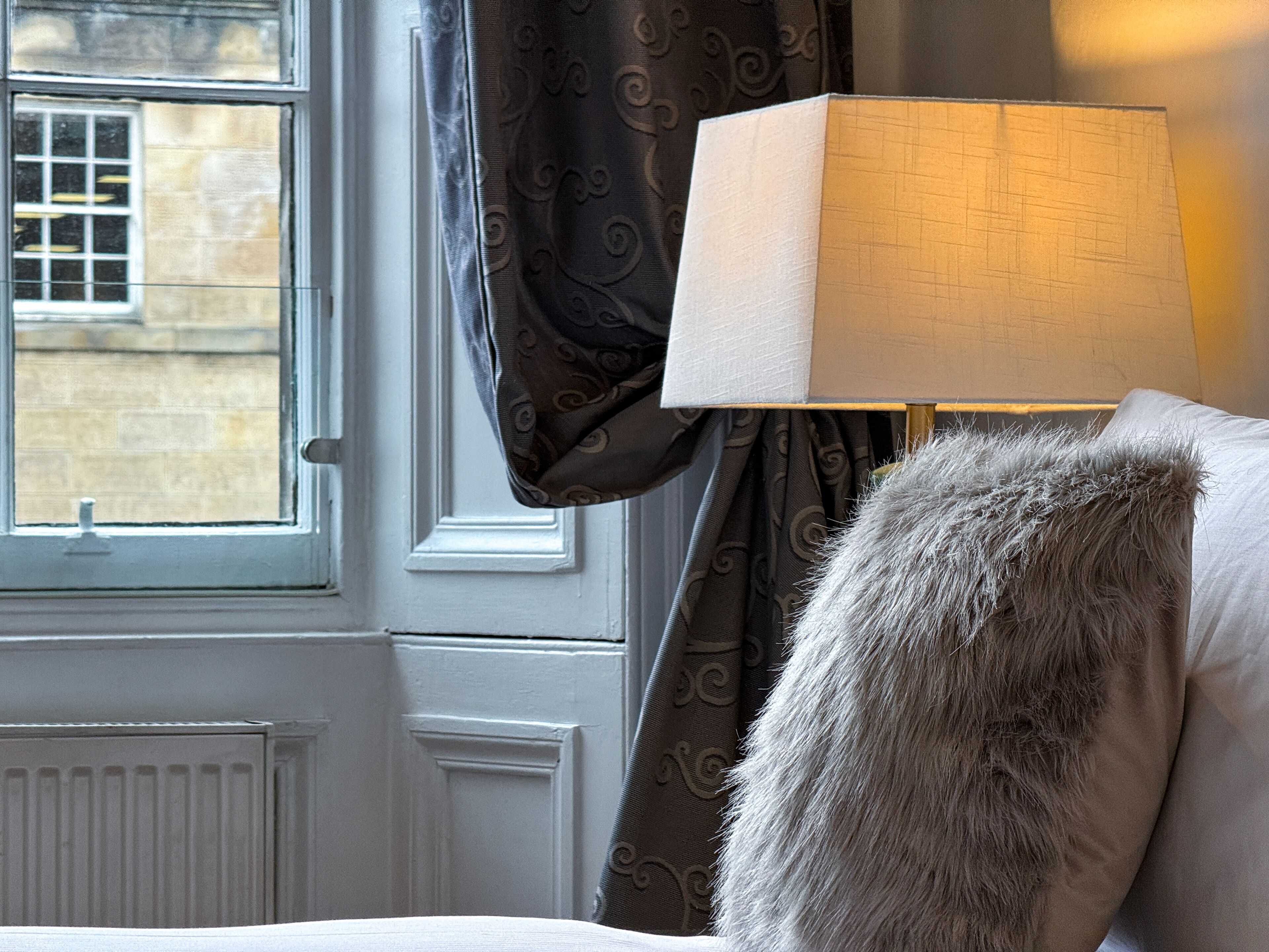 Une pièce avec une fenêtre, une lampe et un plaid en fourrure sur le lit.