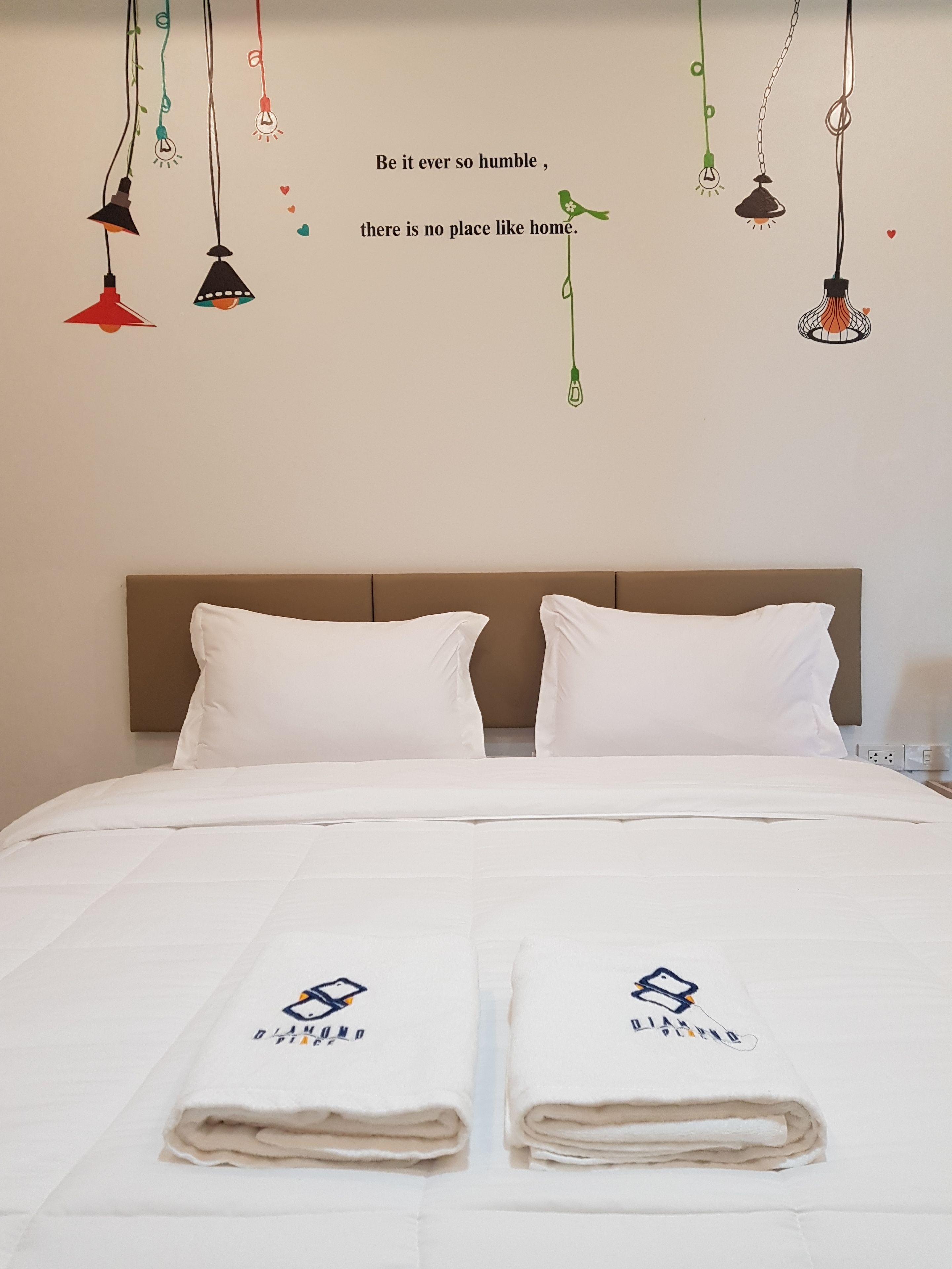A neatly made bed with white linens and pillows, two folded towels with a logo, and a wall with hanging light fixtures and a quote.