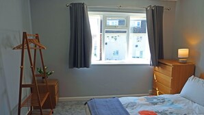 A bedroom with a bed, a wooden dresser, a nightstand, a window with curtains, and a wooden hanger stand. - Birchington Maisonette Near the Sea (Birchington)