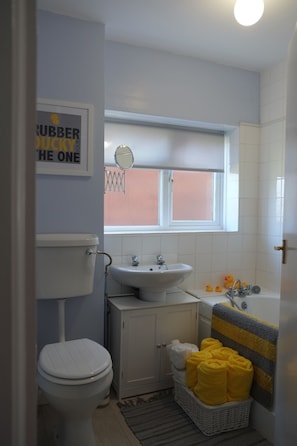 A small bathroom with a toilet, sink, and bathtub. There is a window with blinds, a towel rack with yellow towels, and a framed sign on the wall.