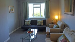 A living room with a sofa, armchairs, a glass coffee table with fruit, a window with curtains, and a wall-mounted lamp. - Birchington Maisonette Near the Sea (Birchington)