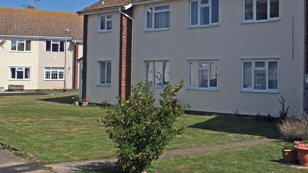 A two-story residential building with a tiled roof, multiple windows, and a small garden area with a bush and potted plants. - Birchington Maisonette Near the Sea (Birchington)