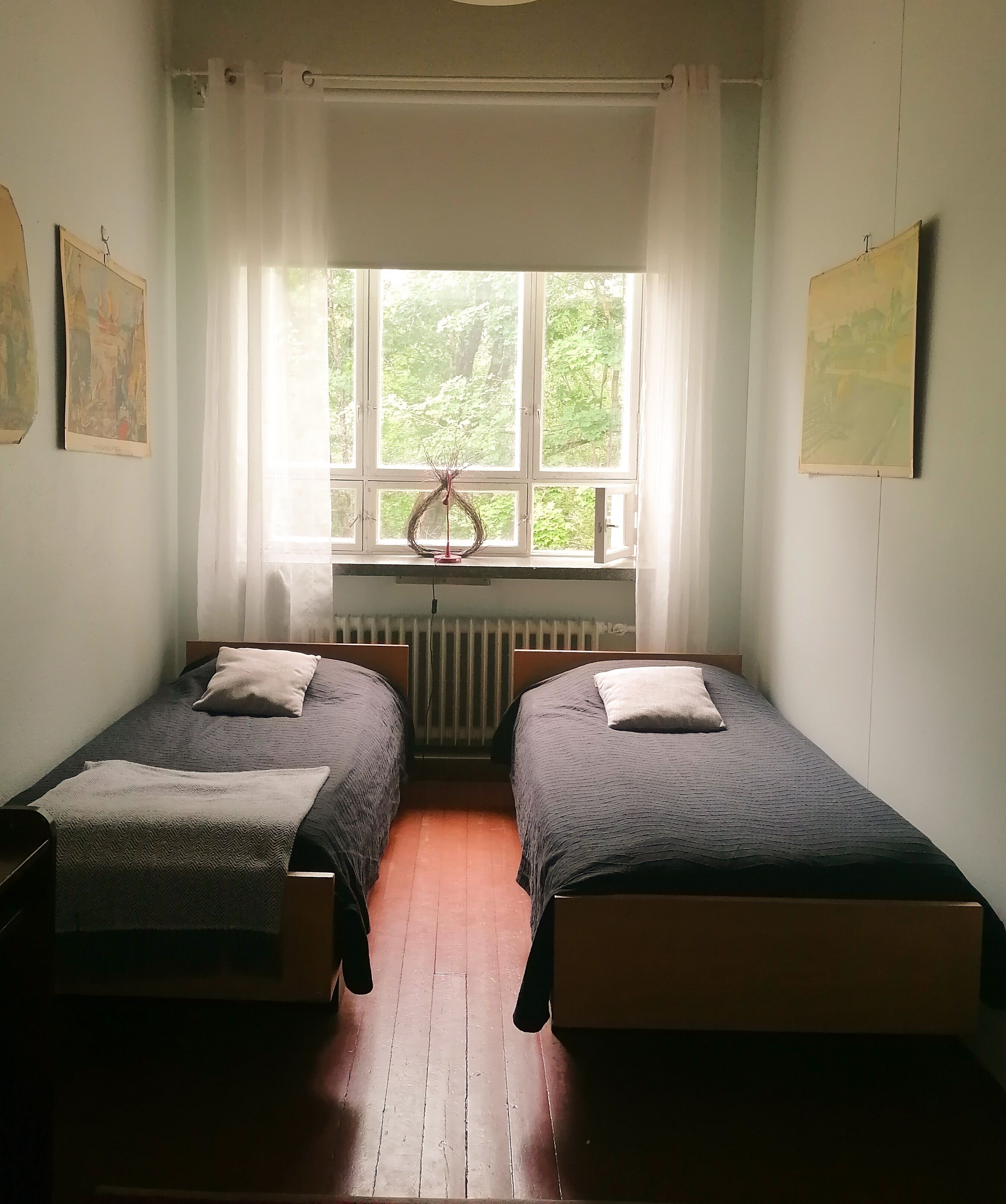 A room with two single beds, a radiator, a window with a view of greenery, and two paintings on the walls.