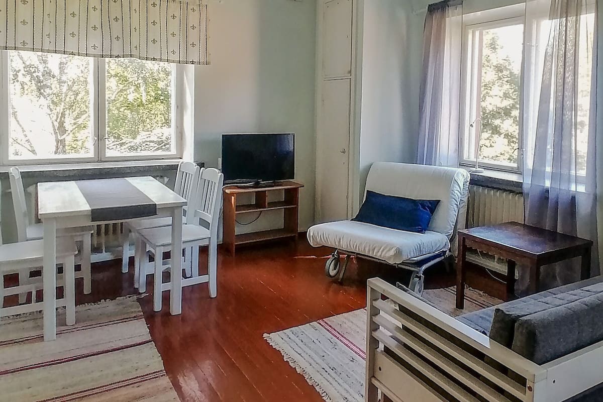 A room with a dining table, chairs, a television, a sofa, and a window with curtains.