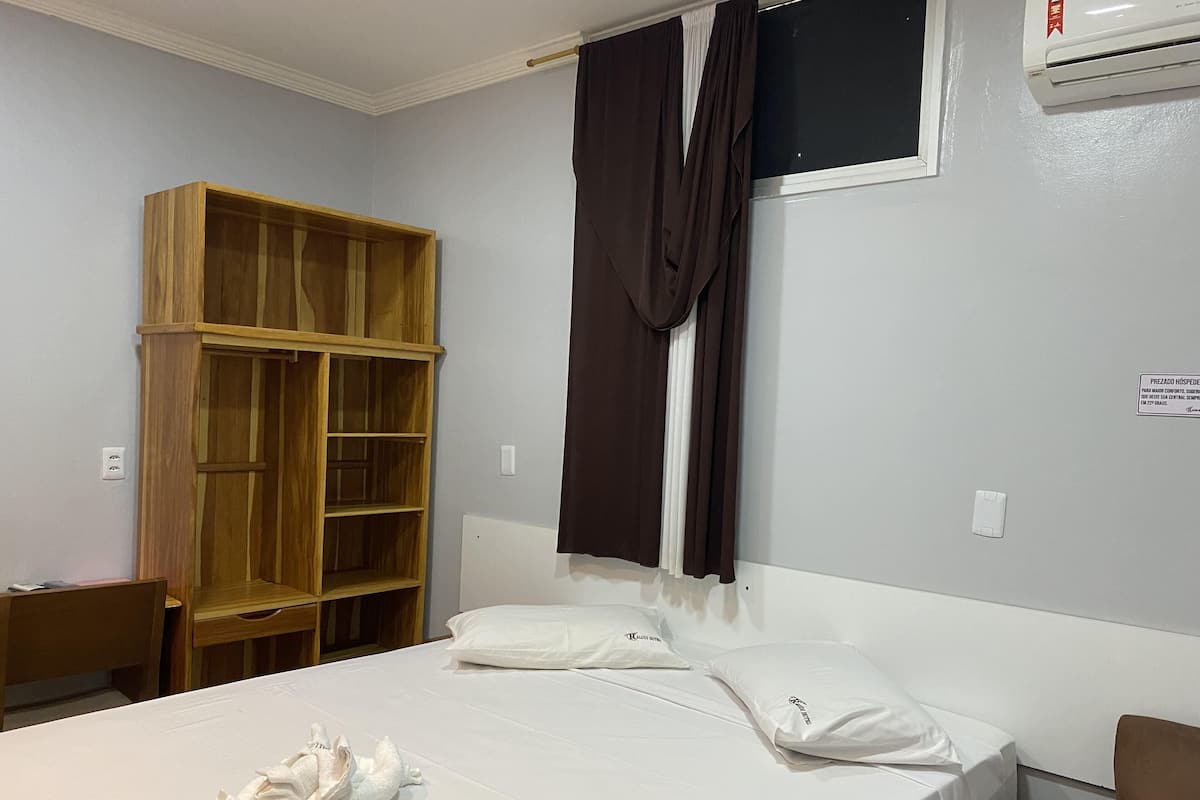 A hotel room with a bed, a wooden wardrobe, a window with curtains, and an air conditioning unit.