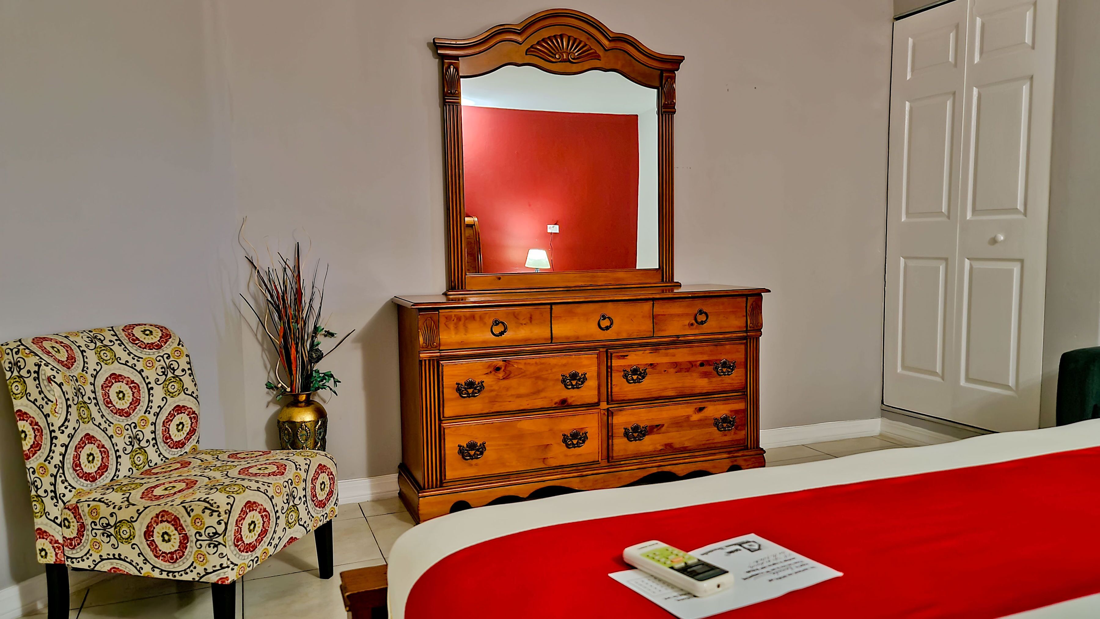 A room with a wooden dresser, a chair, a bed with a red bedspread, and a mirror reflecting a lamp.