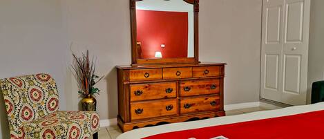 A room with a wooden dresser, a chair, a bed with a red bedspread, and a mirror reflecting a lamp.