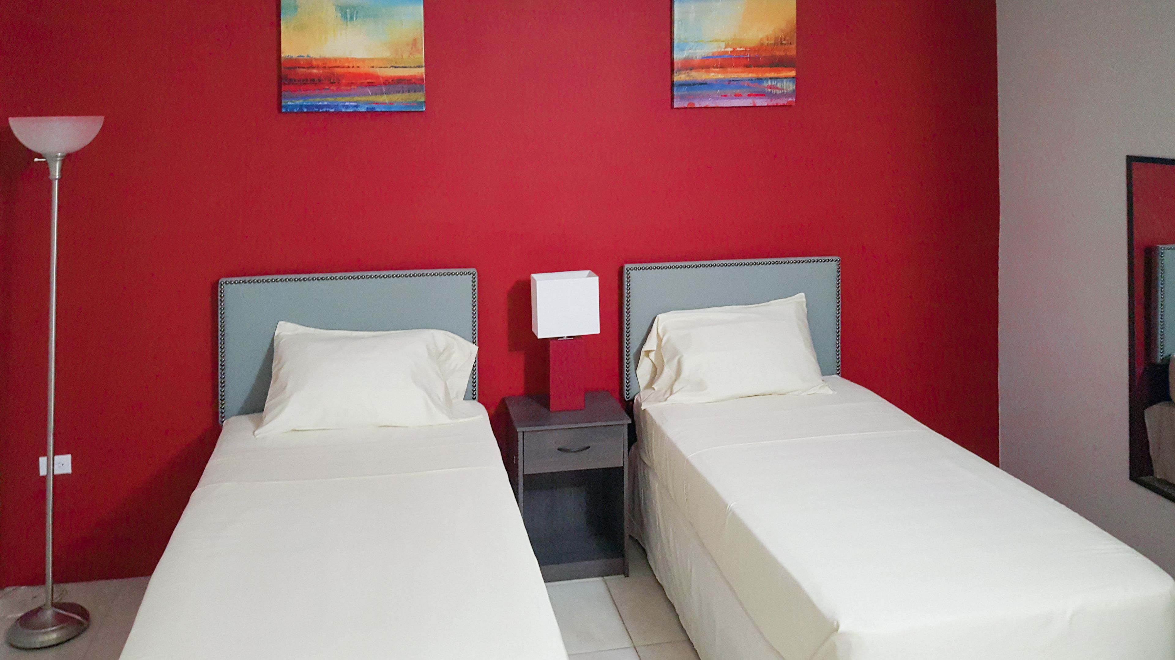 Quarto de hotel com duas camas de solteiro, mesa de cabeceira com abajur e parede vermelha com duas pinturas.