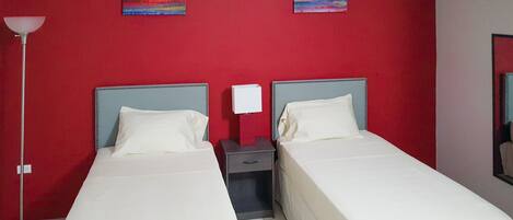 Quarto de hotel com duas camas de solteiro, mesa de cabeceira com abajur e parede vermelha com duas pinturas.