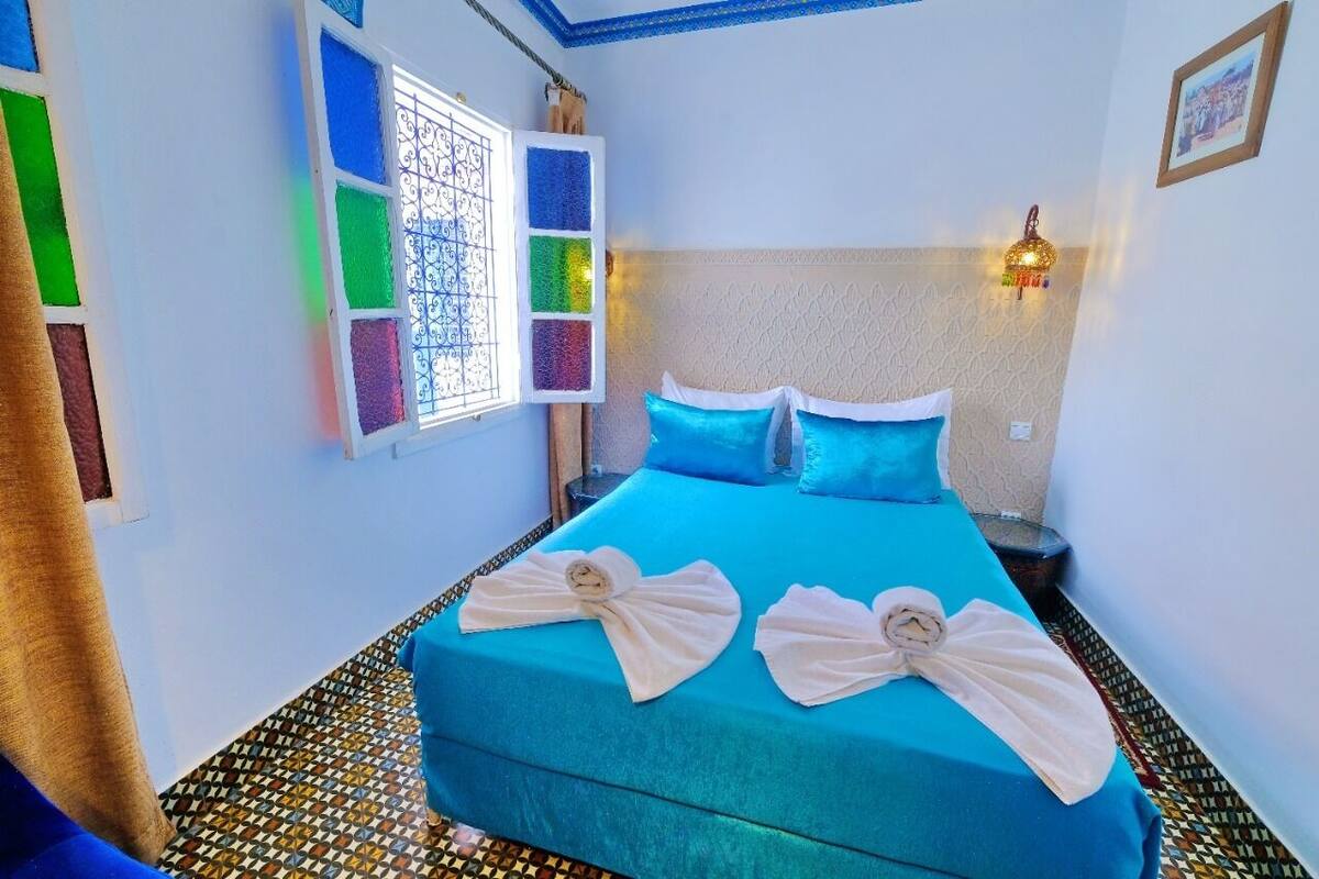 A hotel room with a bed, a window with colorful stained glass, a wall-mounted light fixture, and a patterned floor.
