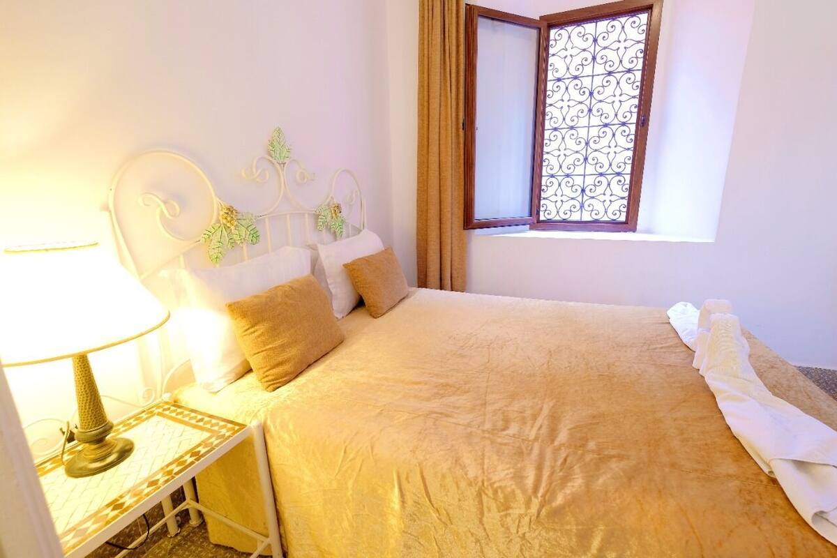 A neatly made bed with a beige bedspread, a white pillow, and a decorative headboard. There is a window with a decorative iron grill and a curtain. A bedside table with a lamp and a patterned tray is visible.