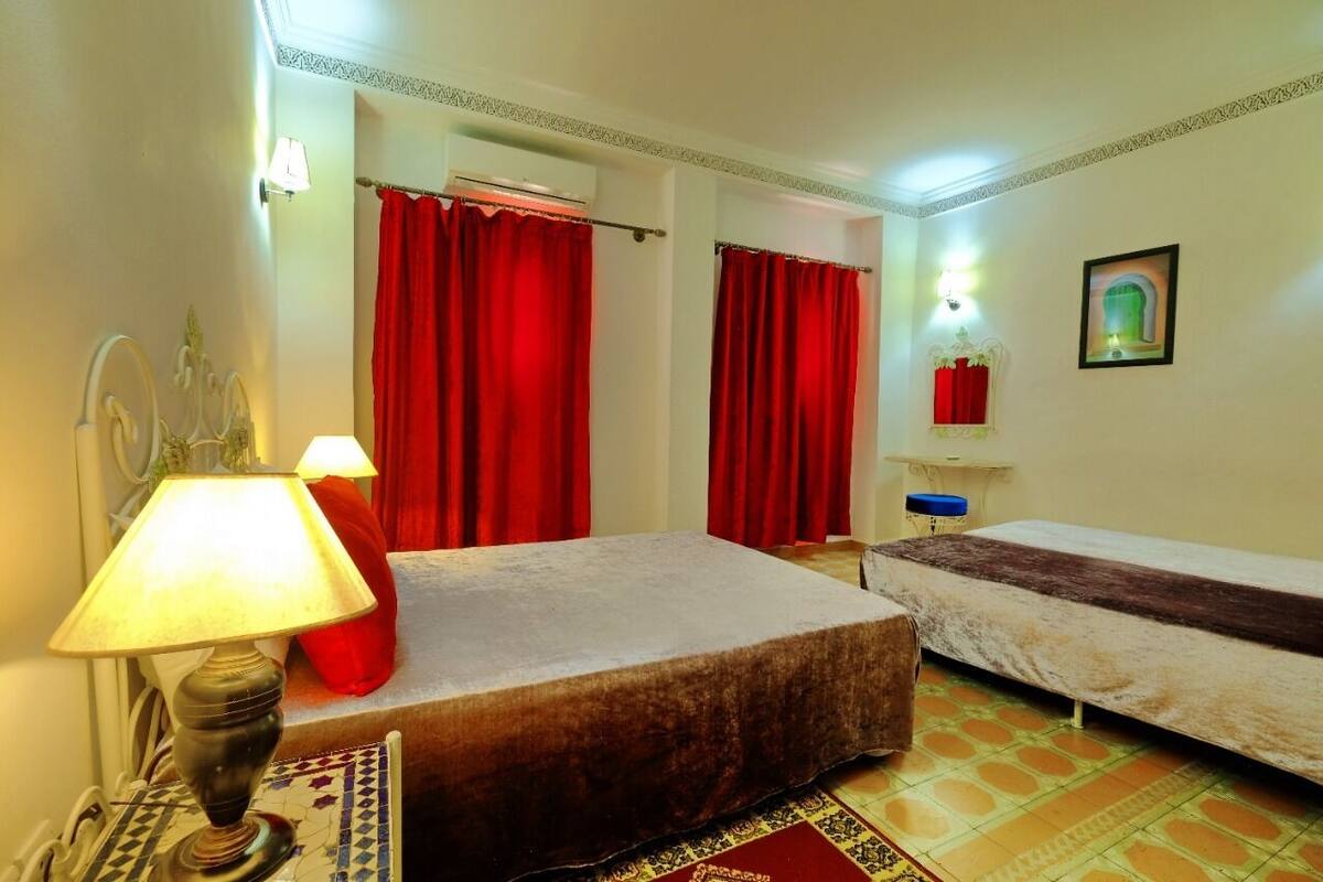 A room with two beds, a red curtain, a lamp, and a small table with a vase.