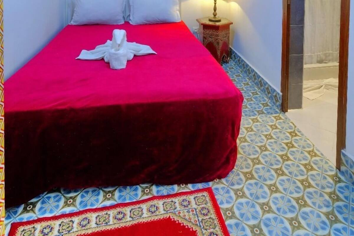 A neatly made bed with a red bedspread and white pillows, a bedside lamp, and a decorative wall piece.