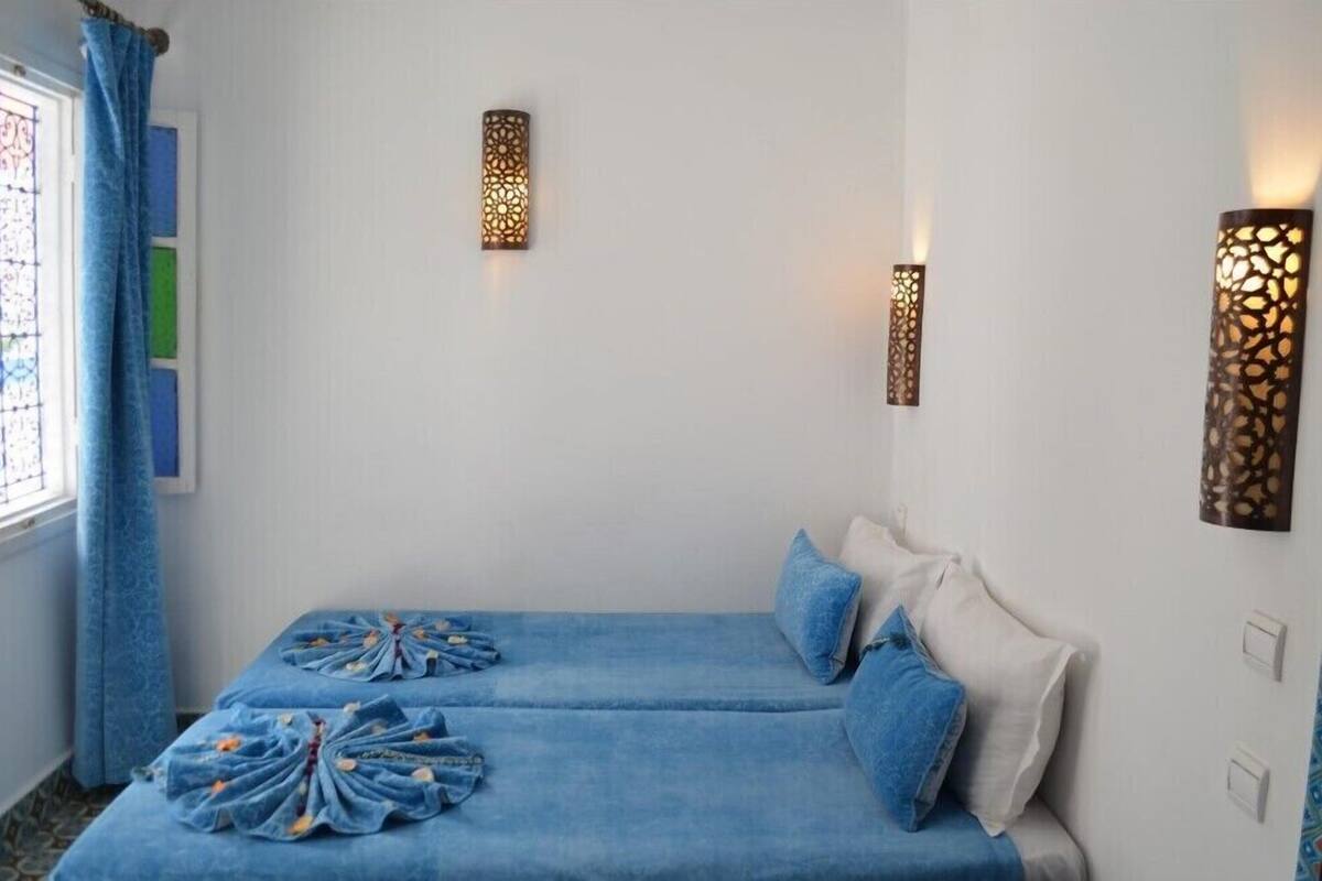 A room with two beds, a window with blue curtains, and wall-mounted lamps.