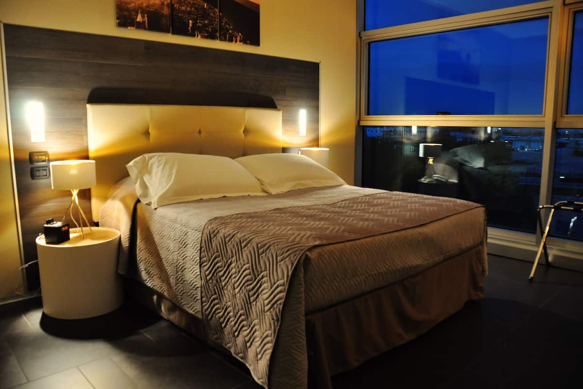 Un dormitorio bien iluminado con una cama grande, una mesita de noche con lámpara y vistas a la ciudad a través de un ventanal.