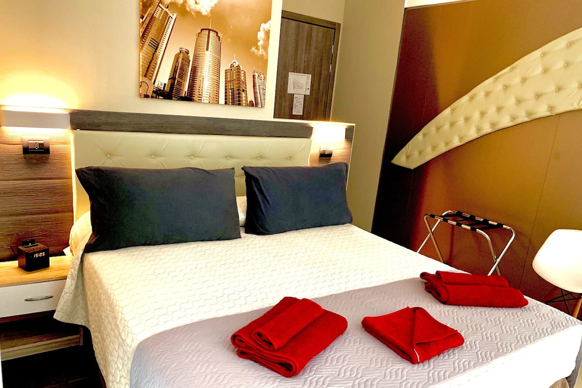Habitación de hotel con una cama grande, dos almohadas, una mesita de noche con un reloj, una obra de arte en la pared y un perchero para toallas.