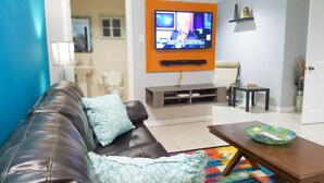 A living room with a leather sofa, a coffee table, a TV mounted on an orange wall, a small table, and a chair.