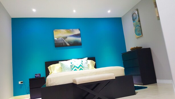 A modern bedroom with a blue accent wall, a bed with patterned pillows, a black nightstand, and a decorative wall piece.
