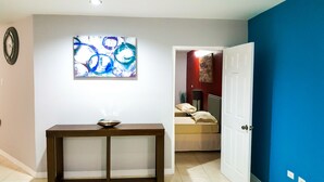A hallway with a painting, a table with a bowl, a bed, and a door.