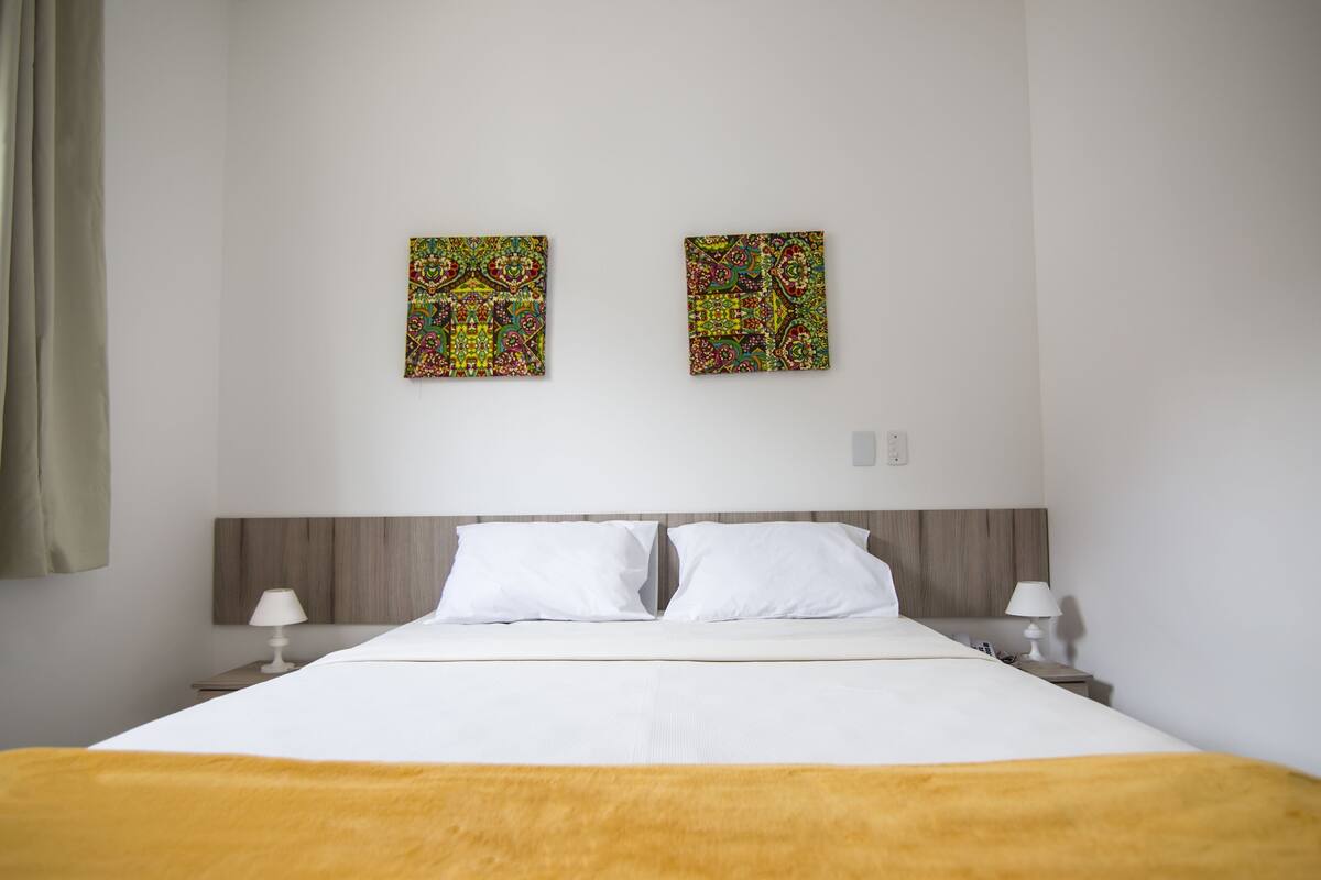 Habitación de hotel con una cama, dos almohadas blancas, un cabecero de madera, dos obras de arte colgadas en la pared, una mesita de noche con lámpara y una cortina a la izquierda.