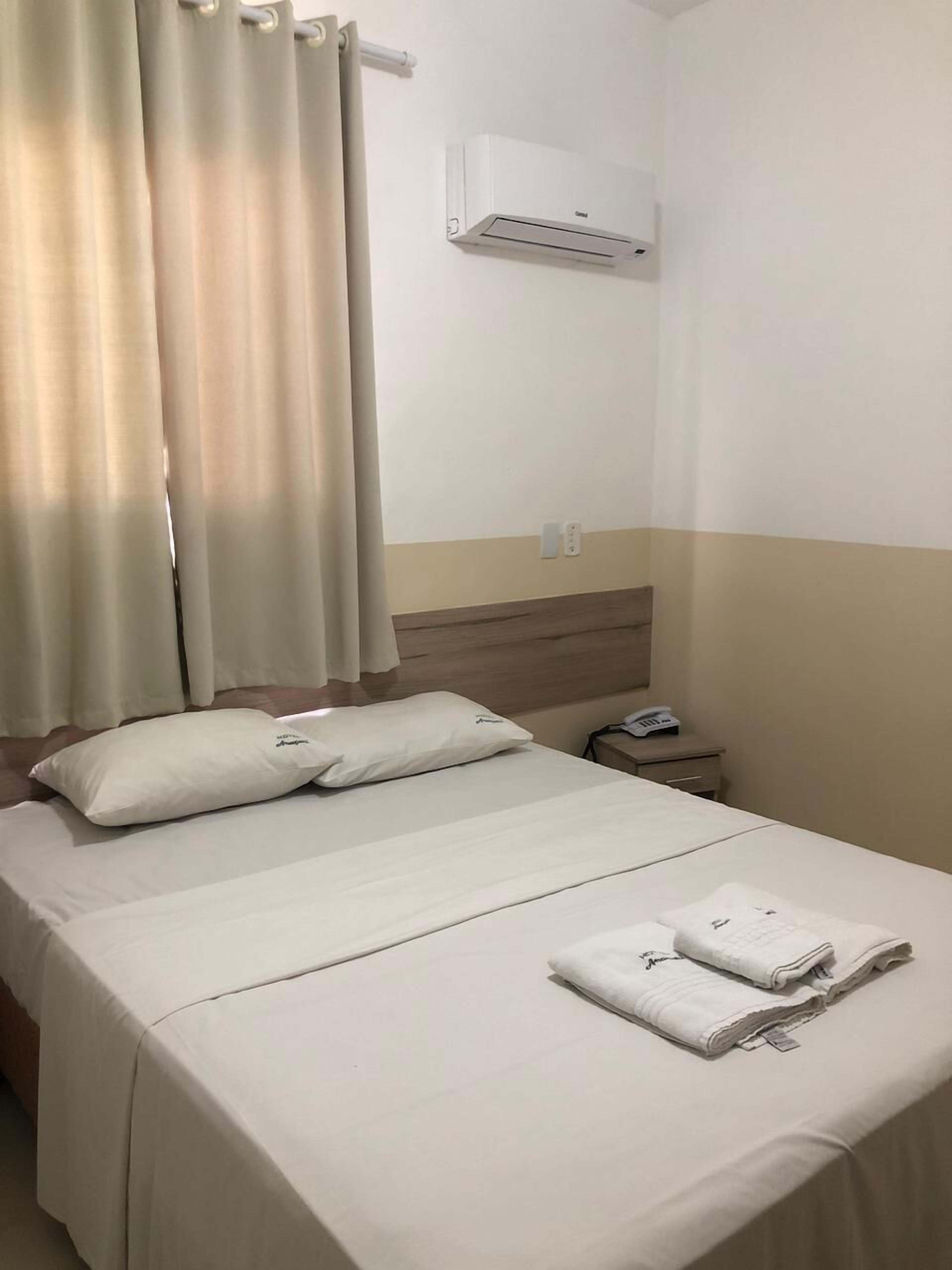 Hotellihuone, jossa on sänky, yöpöytä, ilmastointilaite ja verho.