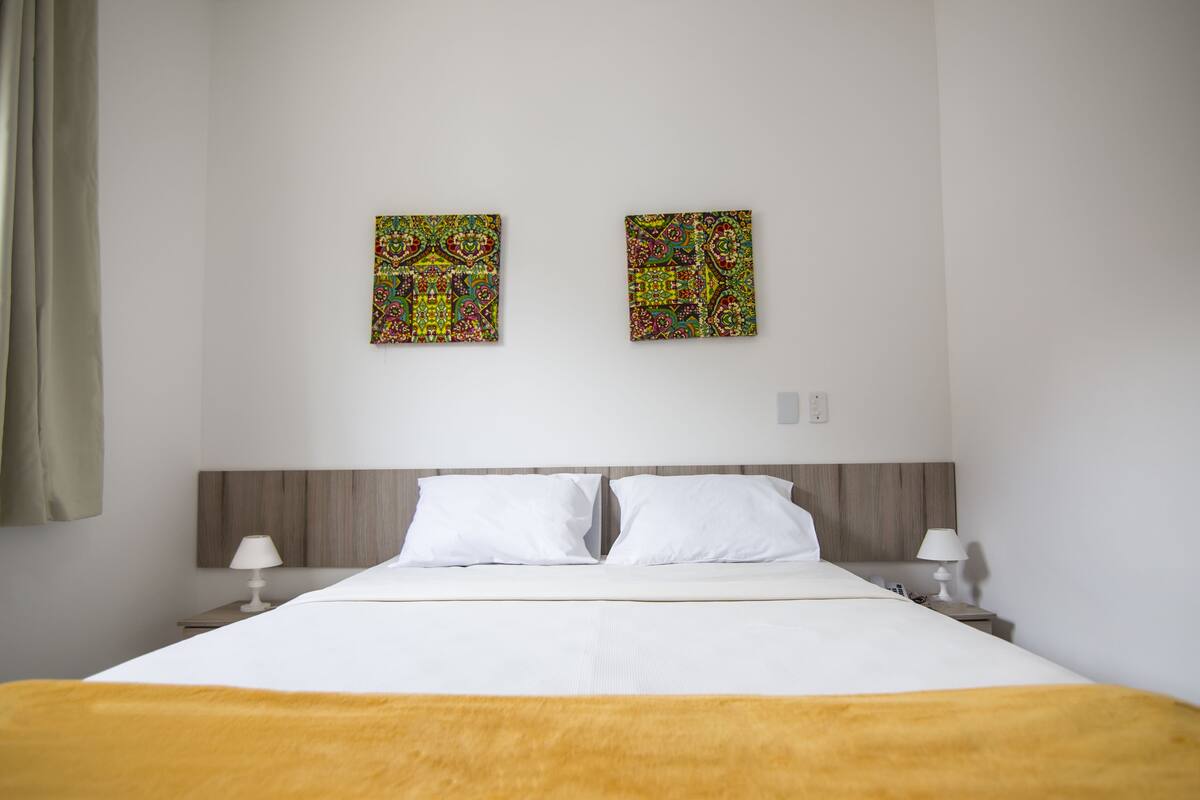 Una habitación de hotel con una cama grande, dos almohadas blancas, un cabecero de madera, dos mesitas de noche con lámparas y dos cuadros abstractos de colores en la pared.