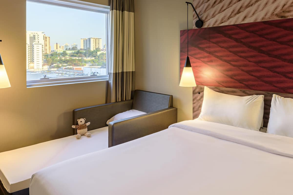 A hotel room with a bed, a chair, a window with a view of buildings, and a wall-mounted lamp.