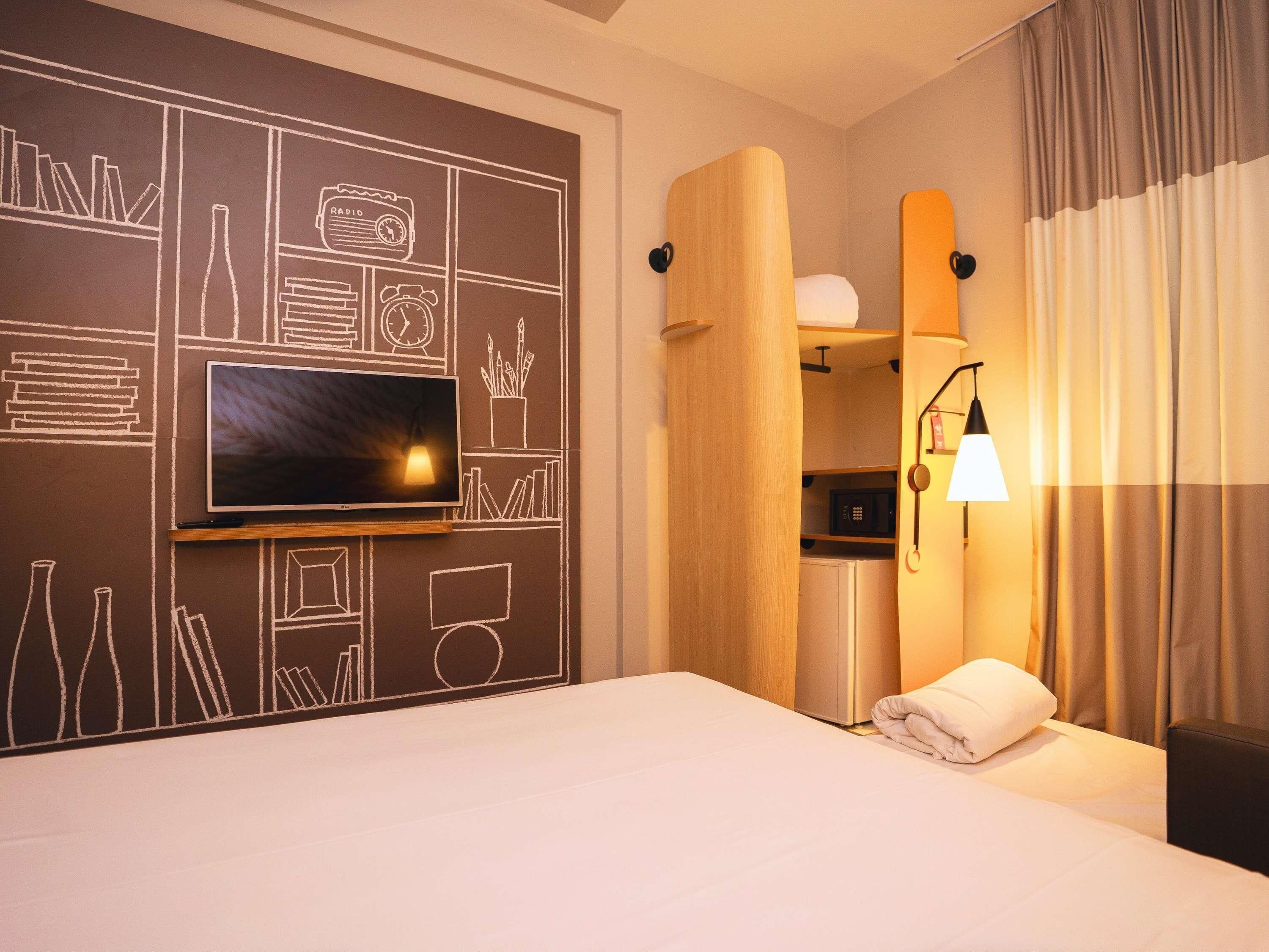 Habitación de hotel con un televisor de pantalla plana grande montado en la pared, una cama con una colcha blanca, una mesita de noche con lámpara y un microondas integrado.