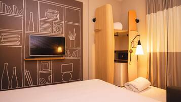 Habitación de hotel con un televisor de pantalla plana grande montado en la pared, una cama con una colcha blanca, una mesita de noche con lámpara y un microondas integrado.