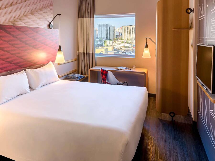Hotellihuone, jossa on suuri sänky, työpöytä, tuoli, televisio ja ikkuna, josta avautuu näkymä kaupunkiin.