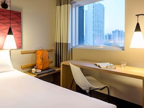 Ein Hotelzimmer mit Bett, Schreibtisch, Stuhl, Fenster mit Stadtblick und einer Pendelleuchte.