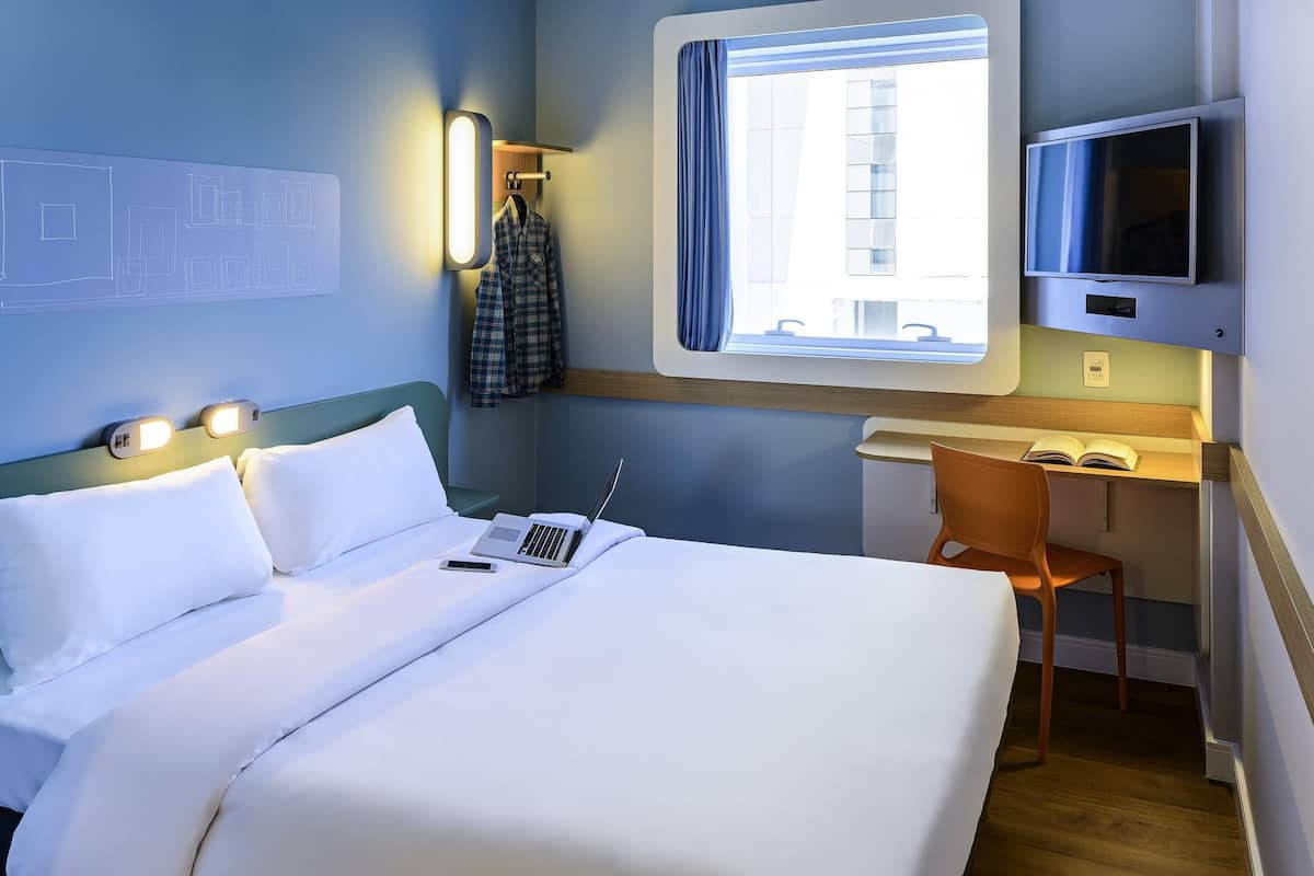 Une chambre d’hôtel équipée d’un lit, d’un bureau, d’une chaise, d’une télévision et d’une fenêtre avec des rideaux.