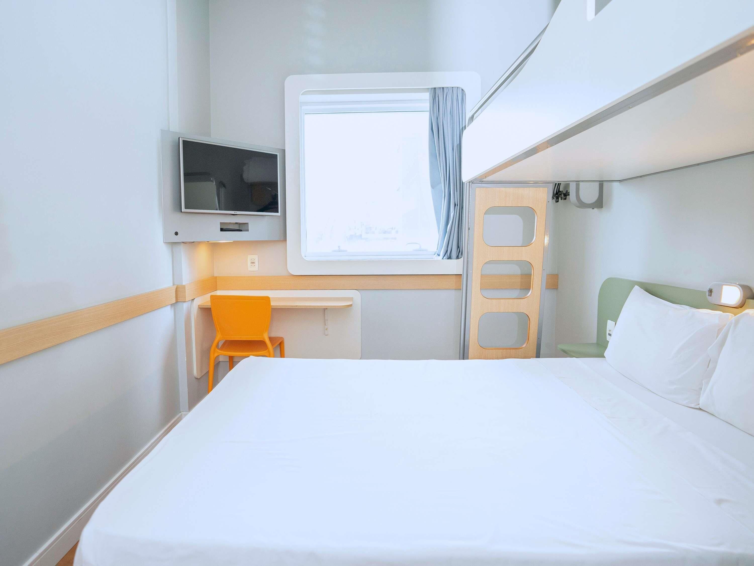 Une chambre d’hôtel de taille réduite, comprenant un lit, un bureau, une chaise, un téléviseur et une fenêtre.