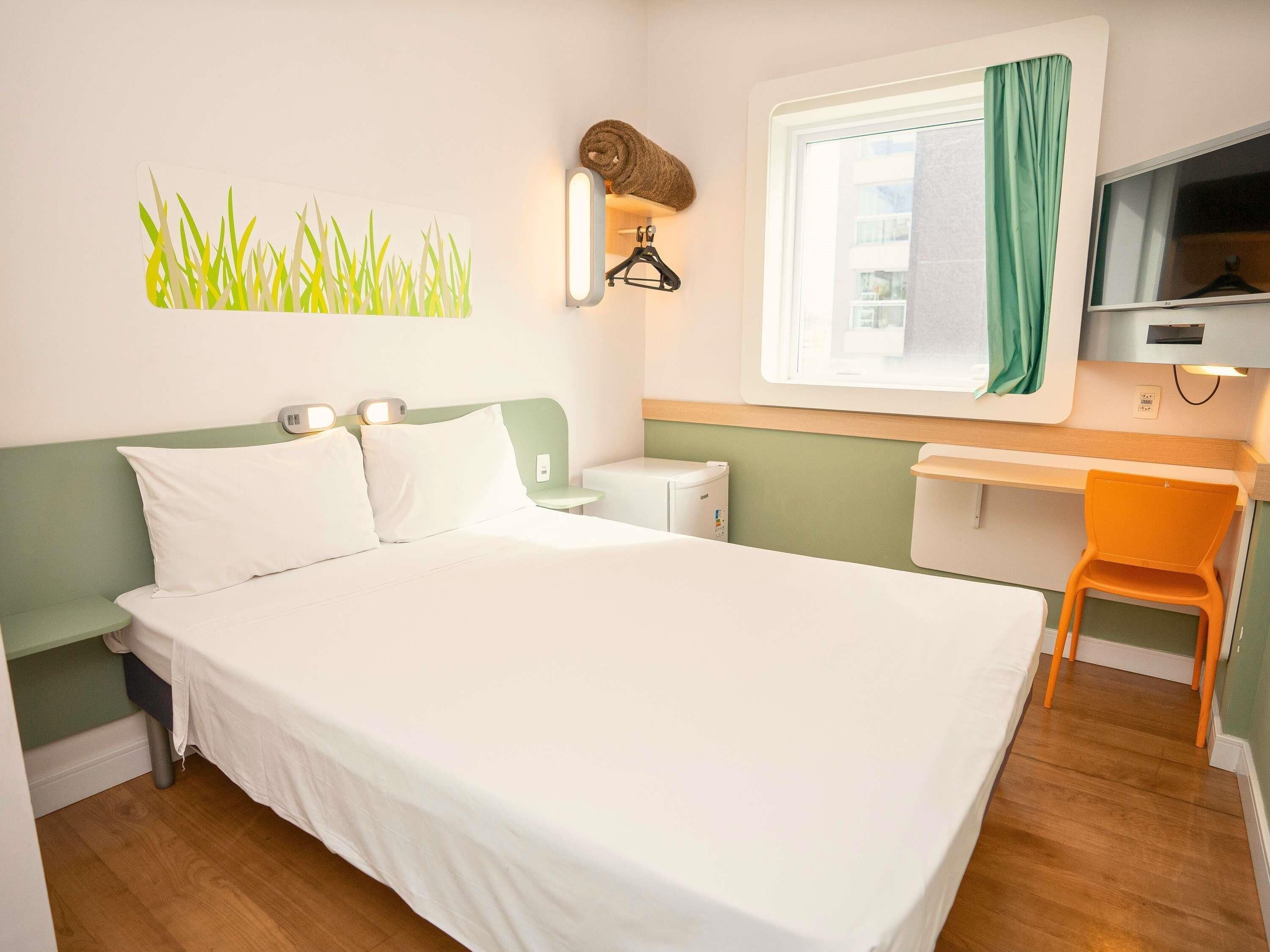 Une chambre d’hôtel comprenant un lit, un bureau, une chaise, une télévision, une fenêtre avec des rideaux et un porte-serviettes fixé au mur.
