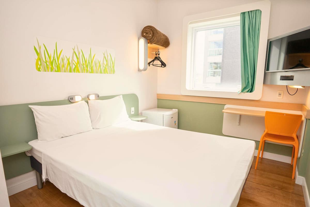 Une chambre d’hôtel comprenant un lit, un bureau, une chaise, une télévision, une fenêtre avec des rideaux et un porte-serviettes fixé au mur.