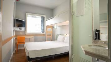 A compact room with a bunk bed, a small desk, a television, and a bathroom with a glass shower enclosure.