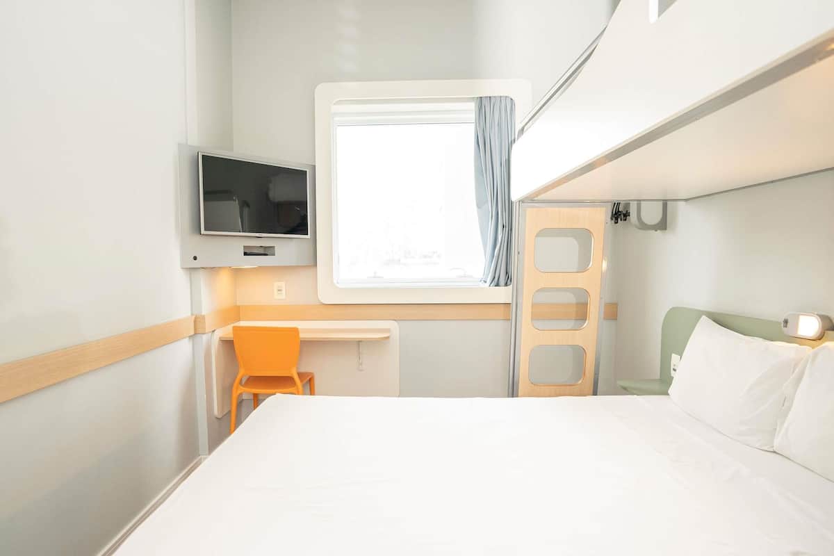 Une chambre d’hôtel de taille modeste, comprenant un lit, un bureau, une chaise, une télévision et une fenêtre avec des rideaux.
