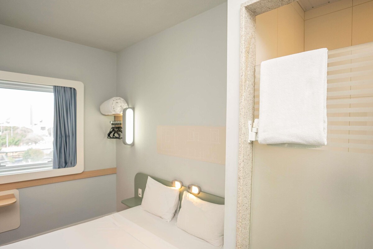 Habitación de hotel con una cama, una ventana con cortinas, una lámpara fijada a la pared y una toalla colgada en una puerta.