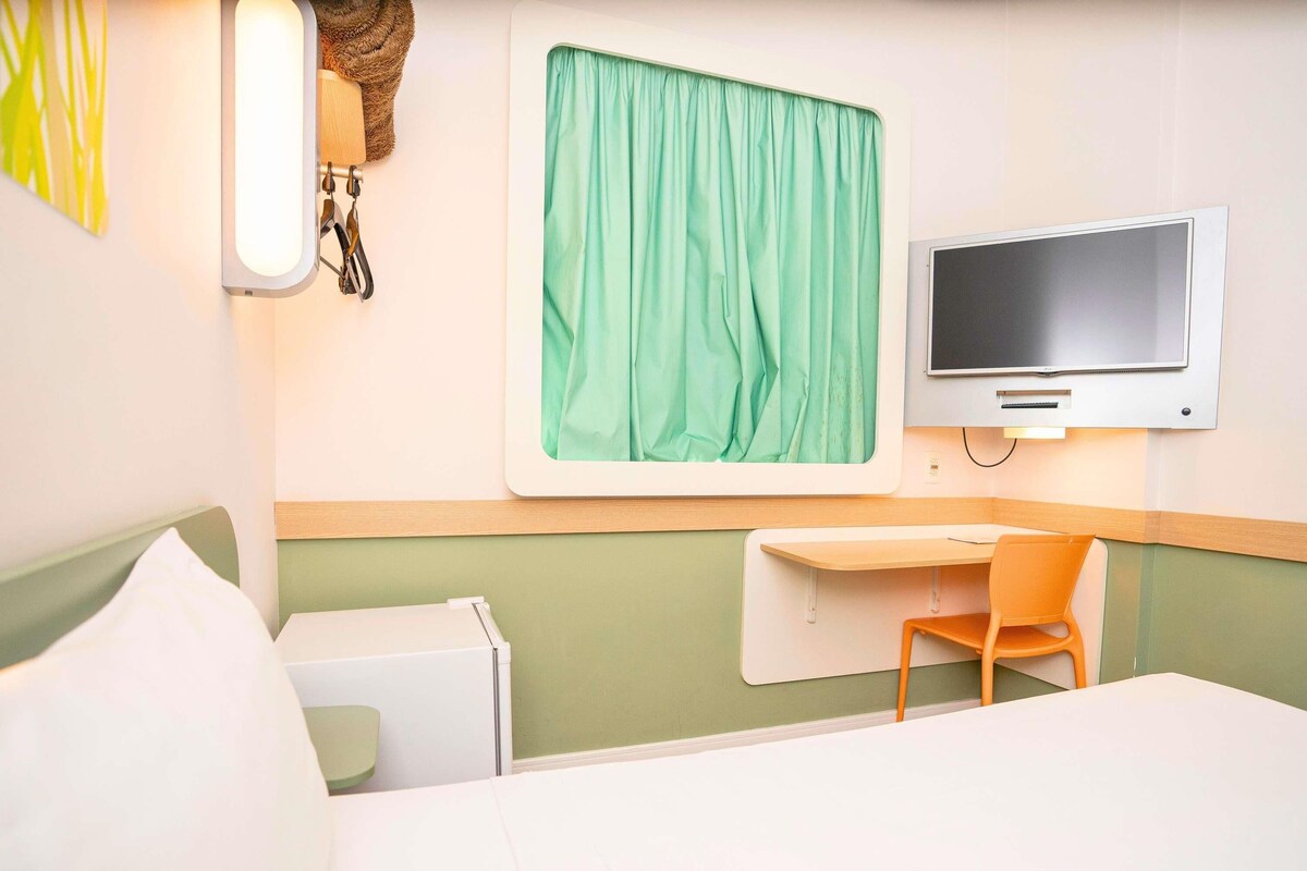 Habitación de hotel con cama, escritorio, silla, televisión y ventana con cortinas.