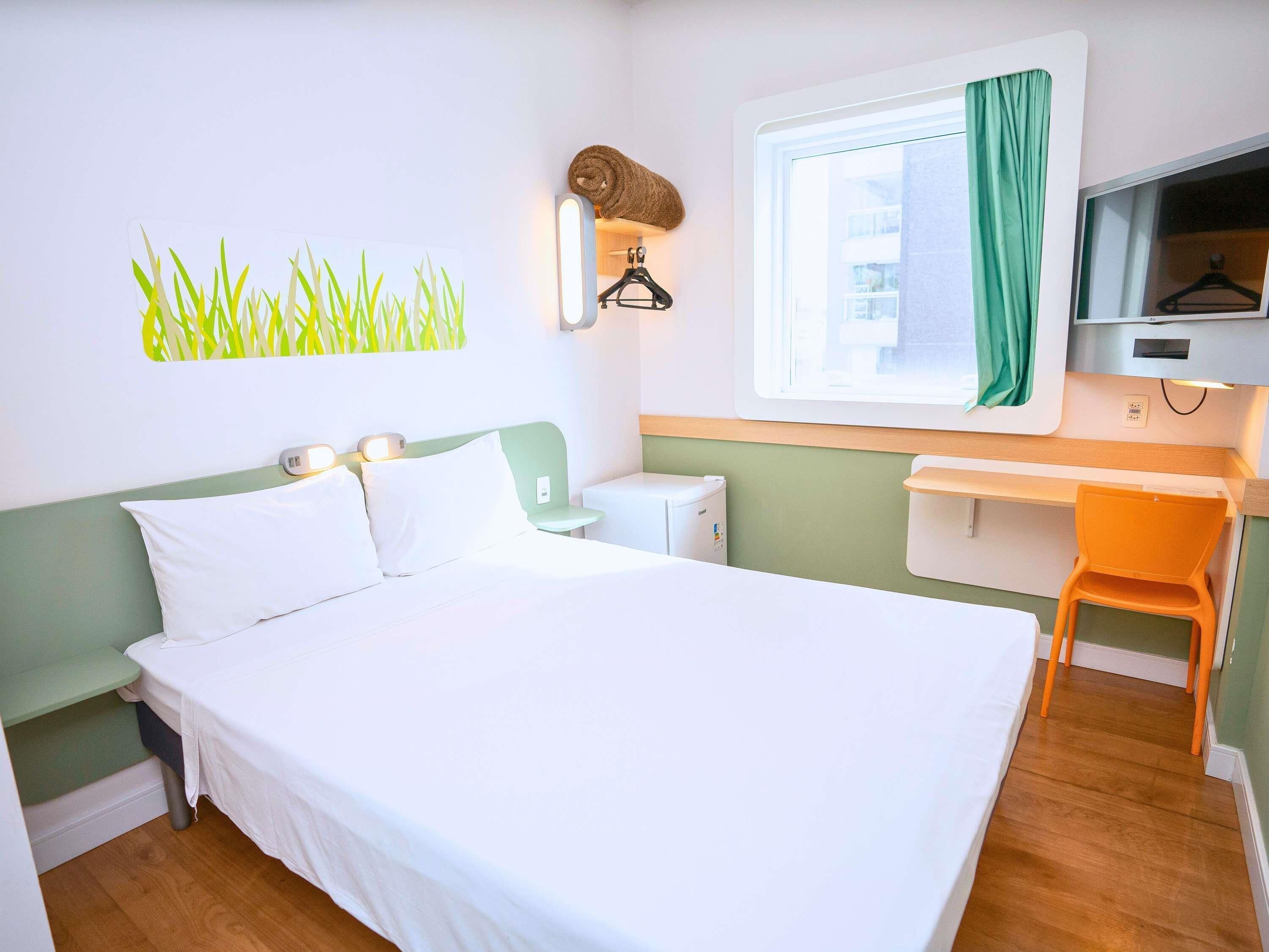 Une chambre d’hôtel équipée d’un lit, d’un bureau, d’une chaise, d’une fenêtre avec des rideaux et d’un téléviseur fixé au mur.