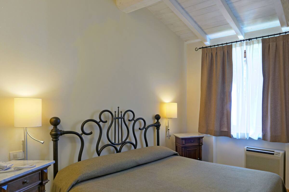A neatly made bed with a headboard, two bedside tables with lamps, a wooden dresser, and a window with curtains.