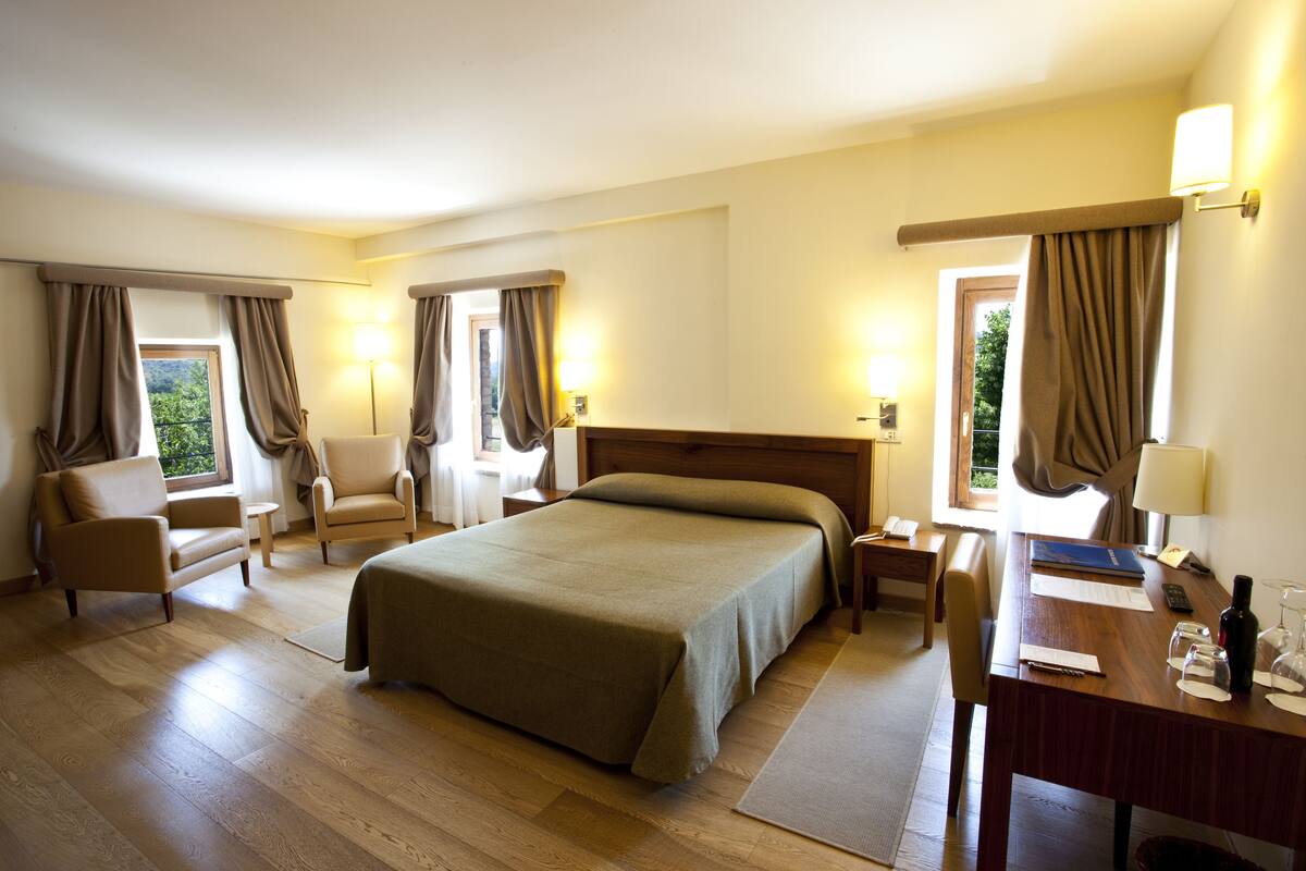 A hotel room with a large bed, two armchairs, a desk with a lamp, and a view of greenery outside.