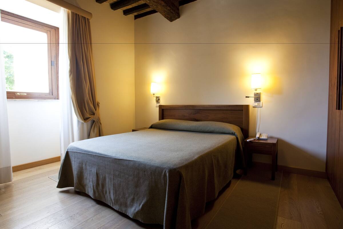 A bedroom with a bed, two bedside lamps, a wooden headboard, a nightstand, and a window with curtains.